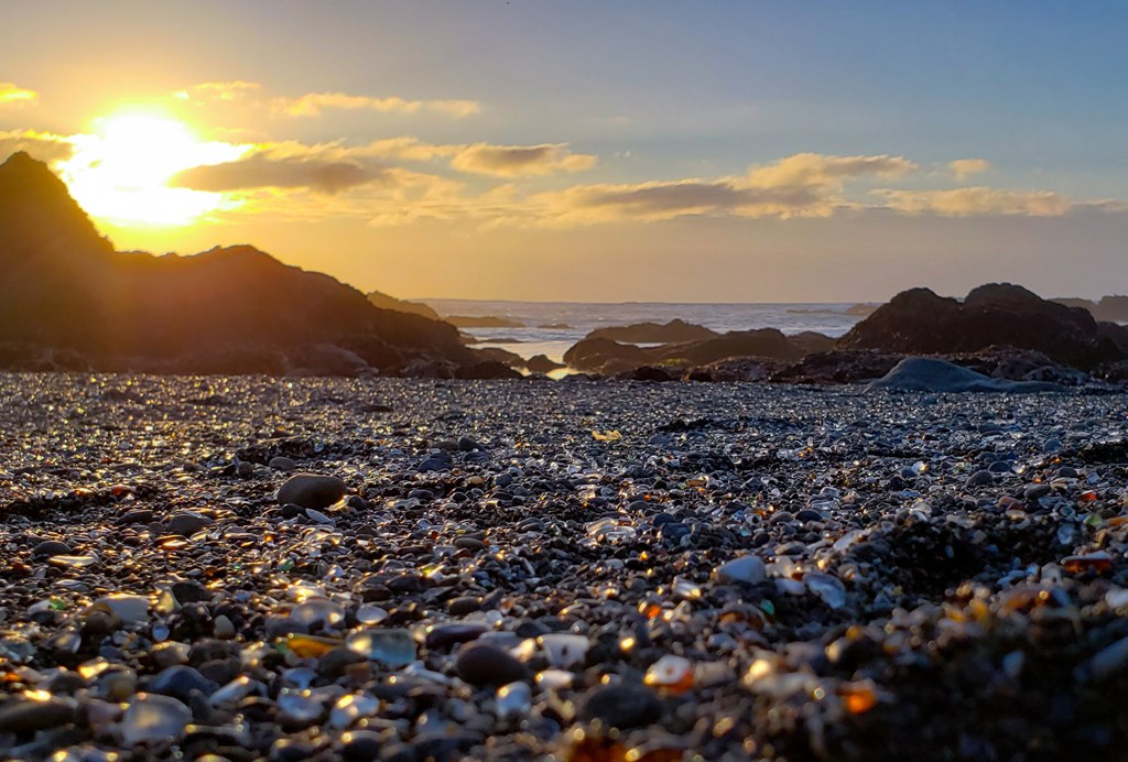 The sun sets over the ocean on a beach covered in sea glass. Fort Bragg Sea Glass Beach, California.