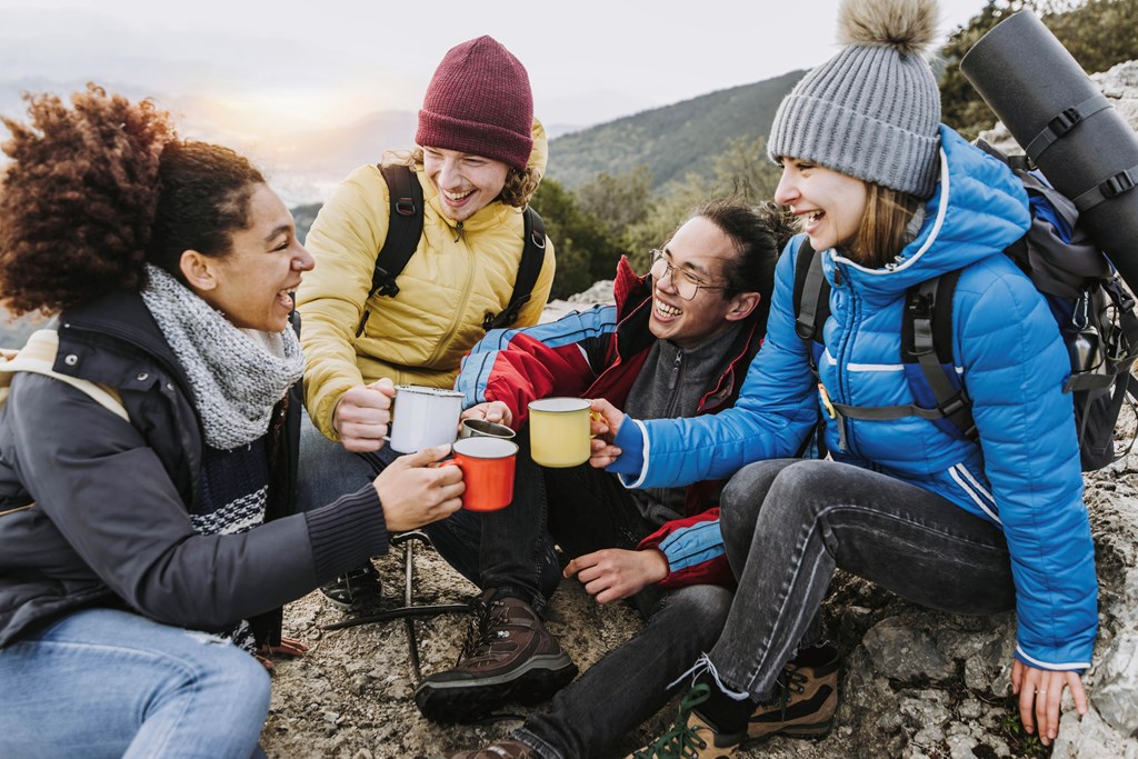 Group of four hikers with backpacks drinking hot coffee on the top of the mountain - Happy friends hiking in nature together.