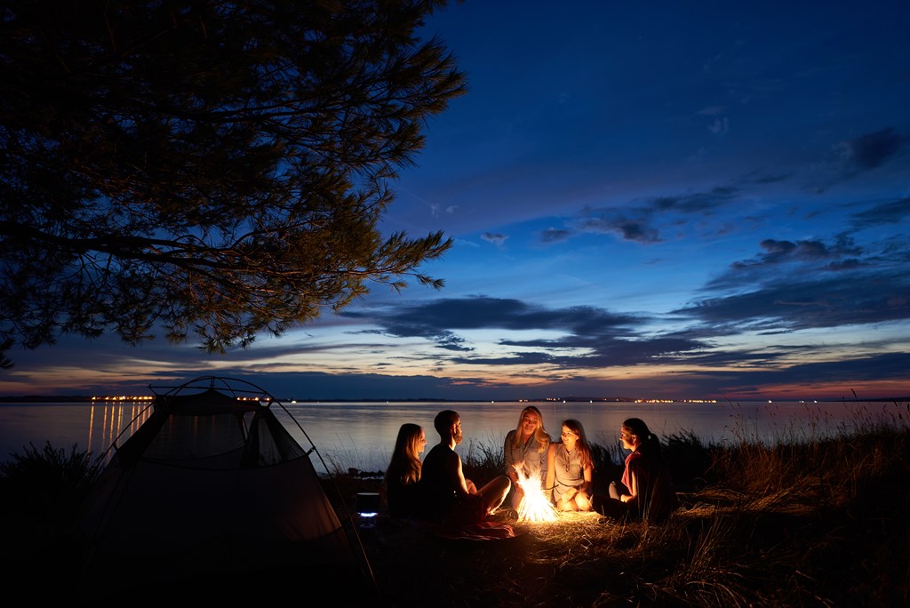Night summer camping on lake shore. Group of five young happy tourists sitting in high grass around bonfire near tent under beautiful blue evening sky.