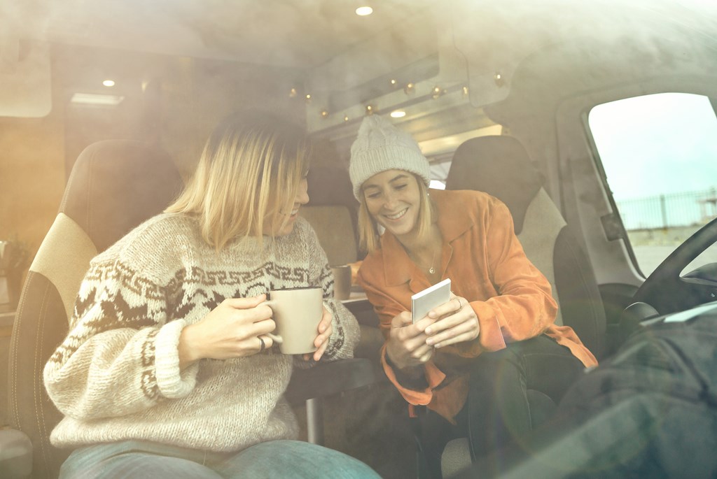 Two women laughing looking mobile with a cup of coffee sitting in the front seat of a camper van.