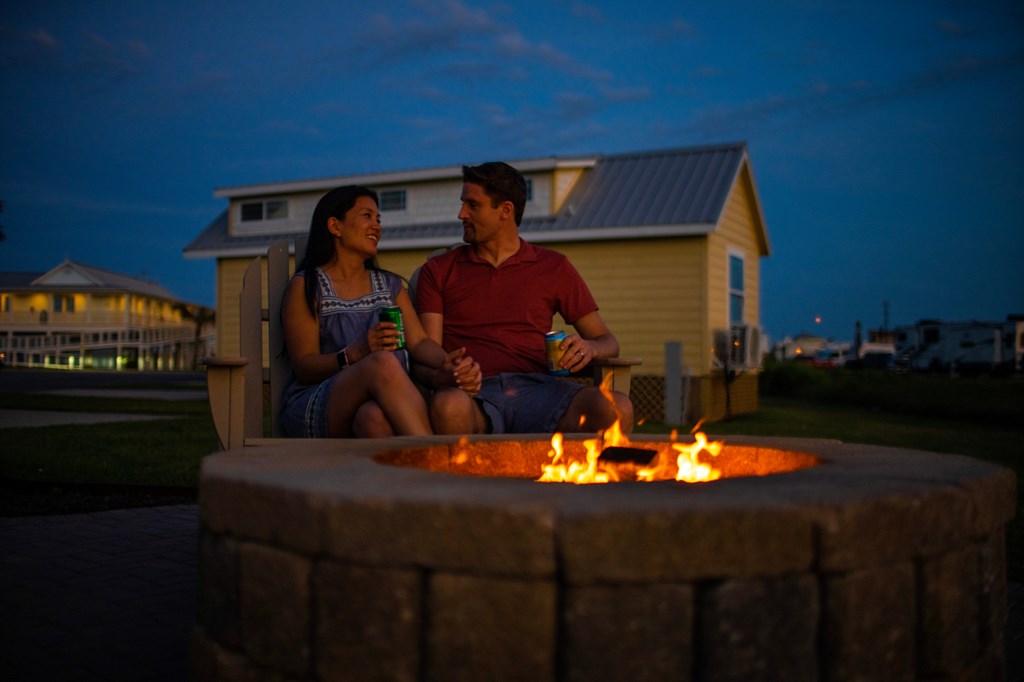 A couple sits at a campfire in front of a cabin at a KOA campground.