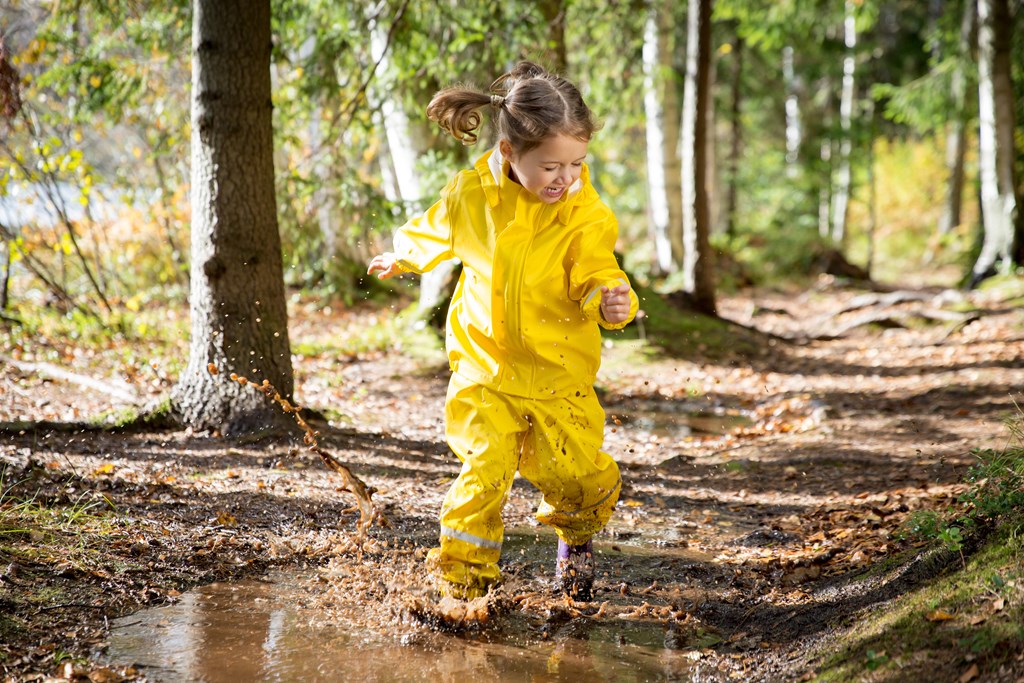Cute little girl jumping in muddy puddle wearing yellow overalls. 