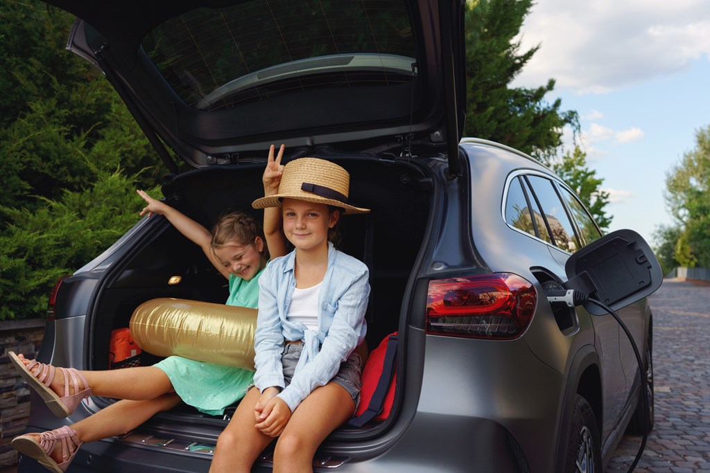 Two girls sitting in the truck of an electric hatchback while waiting for charging car before travelling on summer holiday.