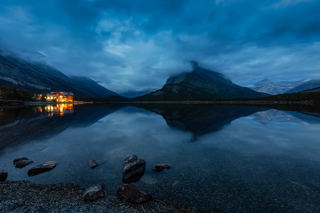 A large lodge at the edge of a mountain lake in the evening.