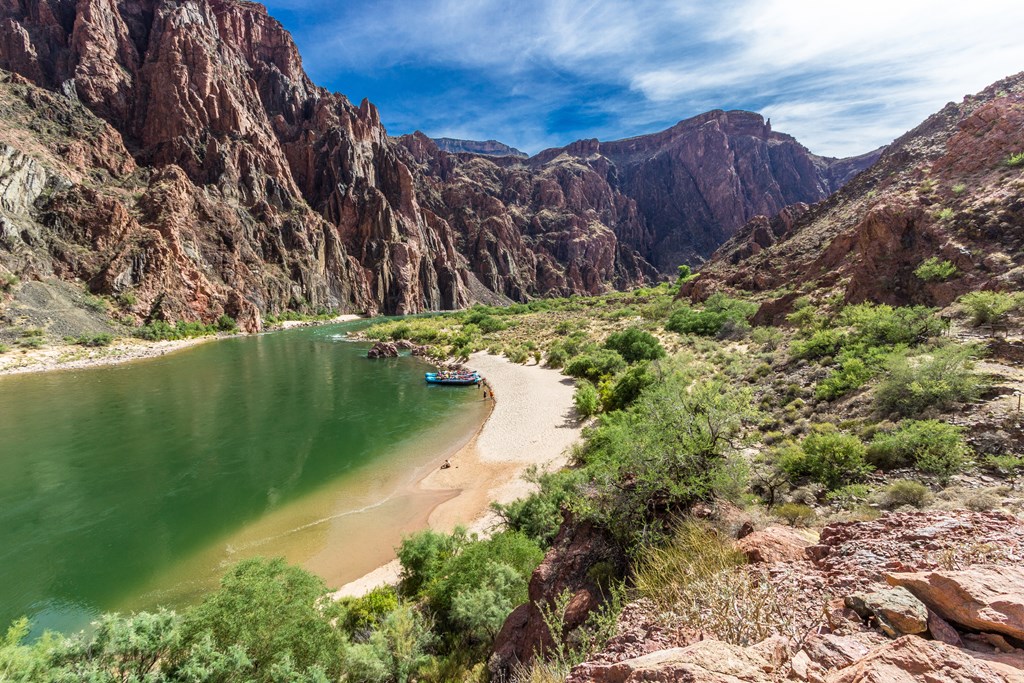 A beach on the Colorado River in the Grand Canyon with rafts and rafters in the distance surrounded by the towering cliffs. At the bottom of the South Kaibab Trail. Nearby is the Black Bridge and Phantom Ranch. The sky is blue with clouds and the river water is green.