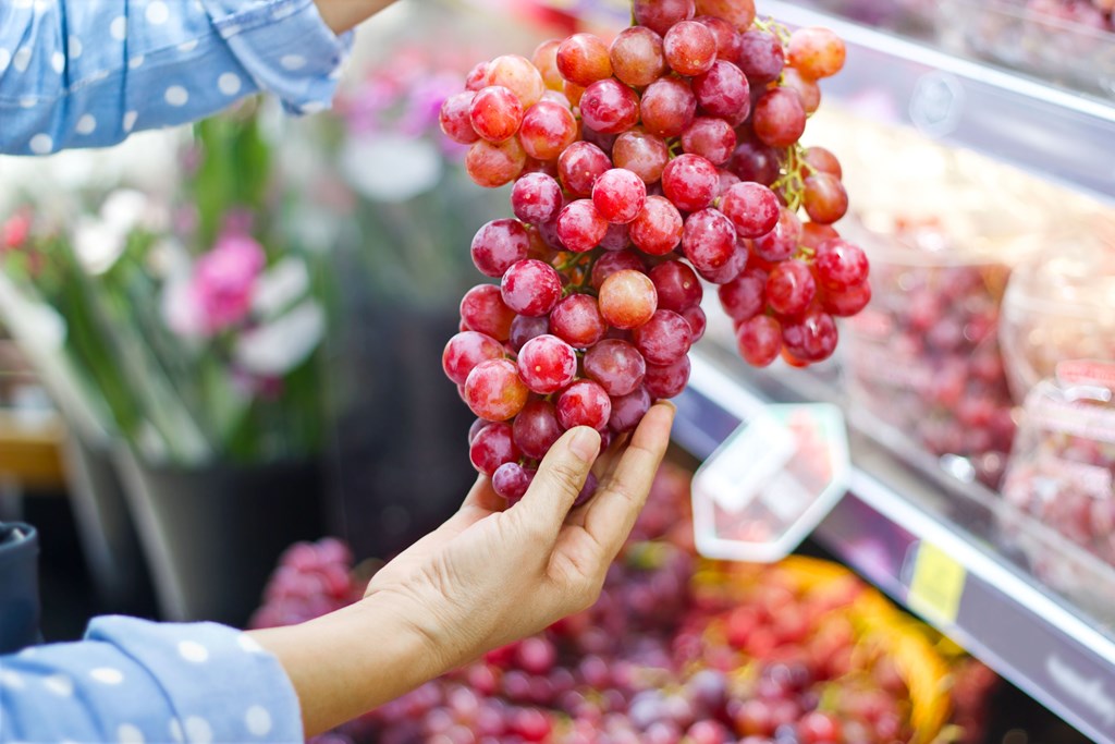 Woman choosing bunch of fresh red grape to buy in supermarket.