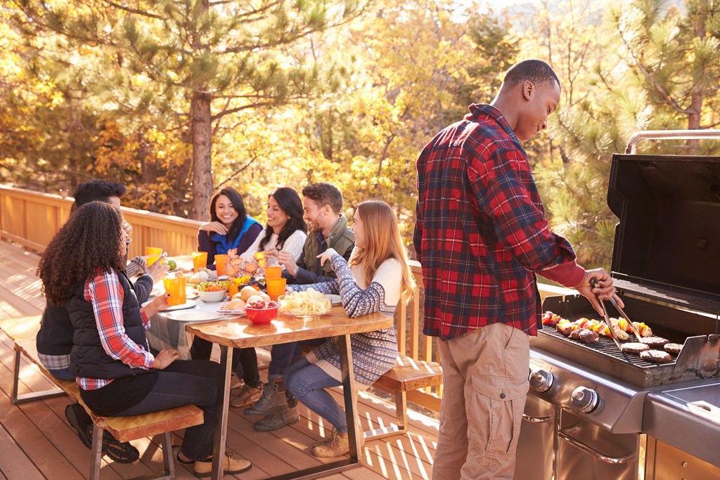 Man barbecues for friends at a table, on a deck in a forest.