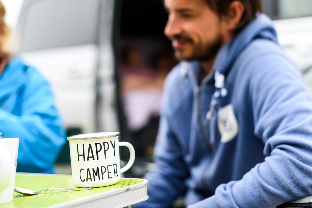 Man camping travelling with breakfast mug and happy camper text. Man drinking coffee or tea from special mug when camping or travelling with motor home or campervan. Vanlife camping concept.