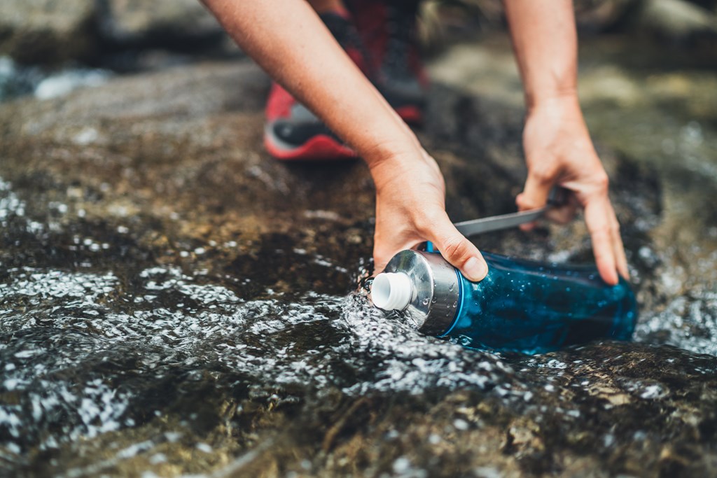 Close-up photo of female hands filling up a fresh cold mountain stream water into a plastic water bottle while on a hiking break. 
