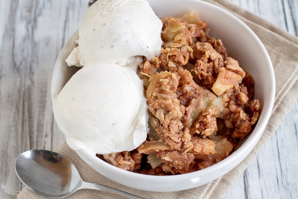 Fresh hot homemade apple crisp or crumble with crunchy streusel topping topped with vanilla bean ice cream over rustic white table. Selective focus with blurred background.