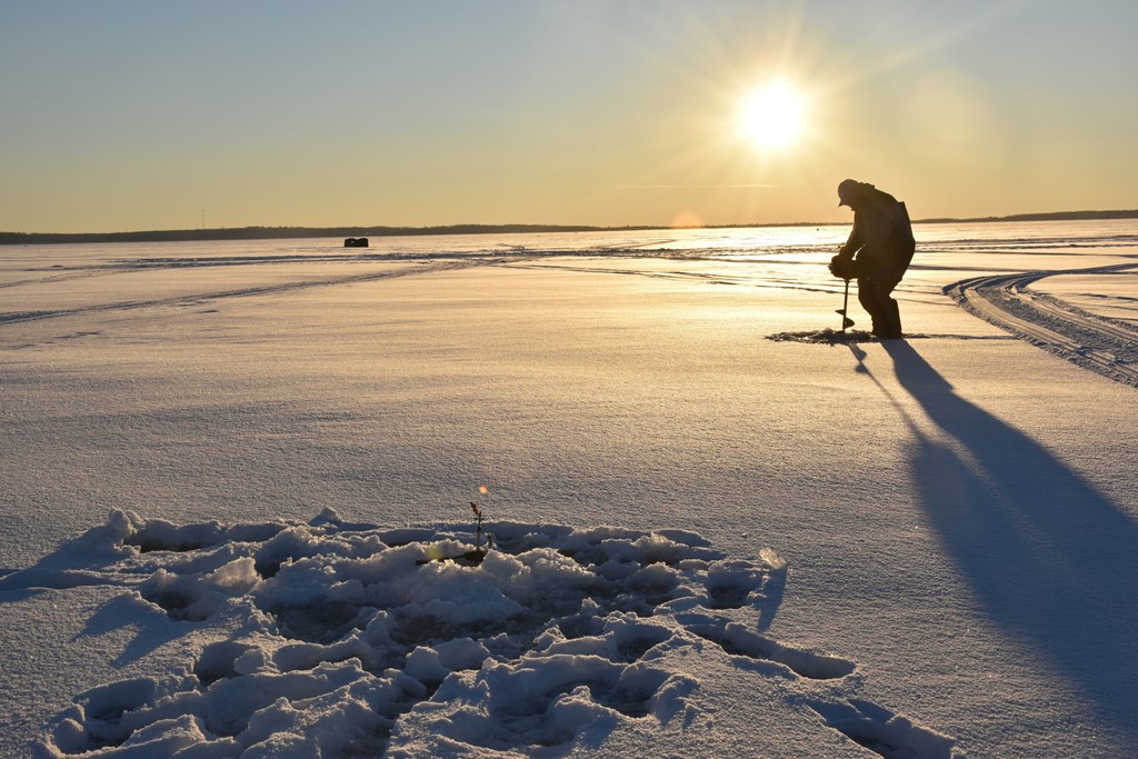 An ice fisherman drilling a hole in the ice and snow with the sun low in the background.