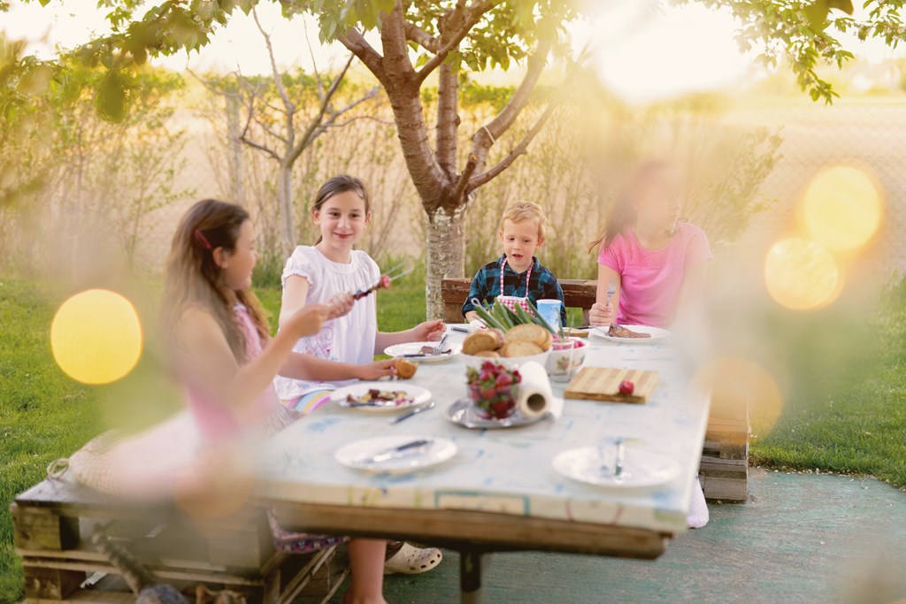 Picture of four kids sitting by the table in nature and eating. 