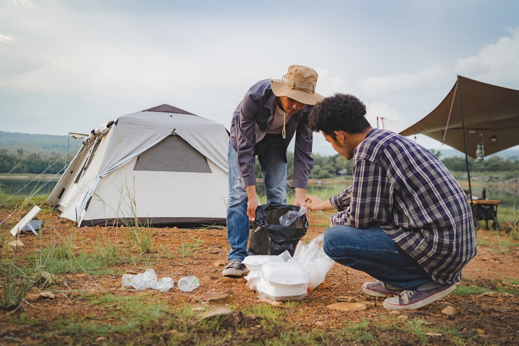 Two young men clean up trash at their campsite with a tent in the background.