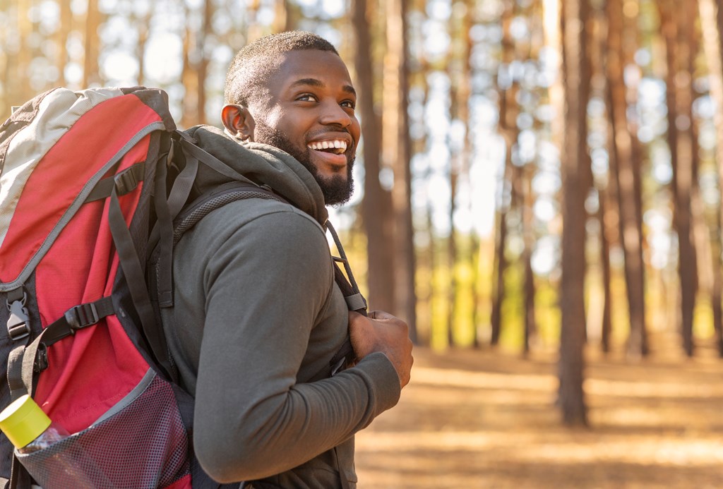 Smiling young  man wearing backpack standing on autumn forest trail, looking aside, hiking alone.