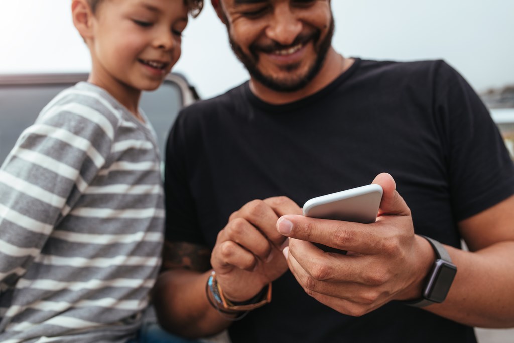 Close up of happy father and son in front of the car looking at the mobile phone. Young man and little boy using smart phone while on road trip.