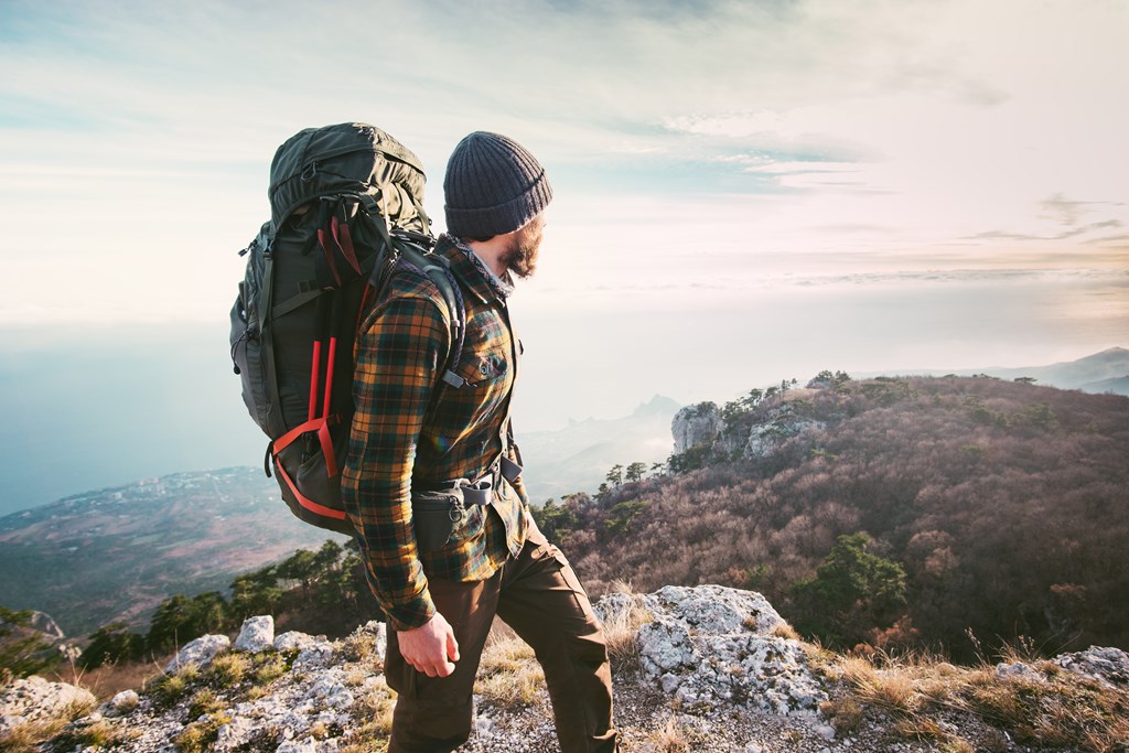 Bearded man in a flannel shirt and beanie wearing a hiking backpack looks on the view from a mountain.