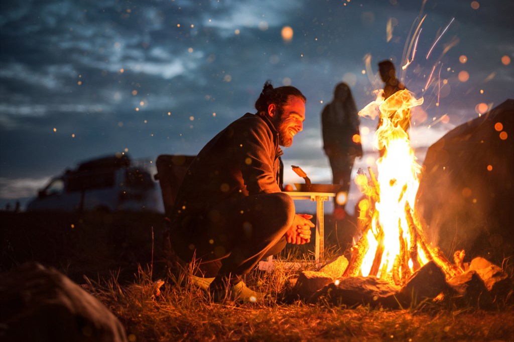 A smiling man in his 20s is crouching in front of a campfire to warm himself. In the background you can see a van and the silhouettes of two other people.