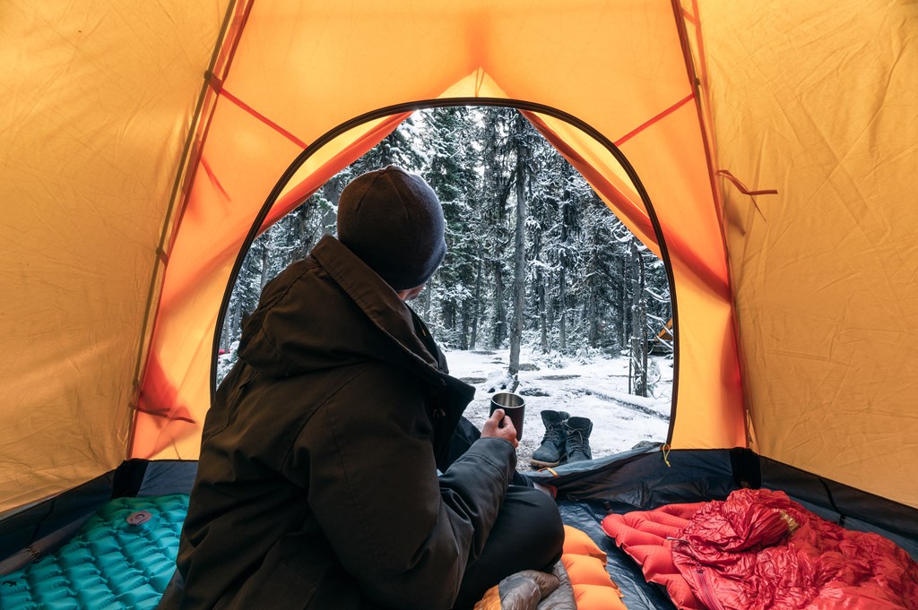 Man traveler wearing winter coat with sitting and holding a coffee cup in orange tent on campsite in the winter snow.