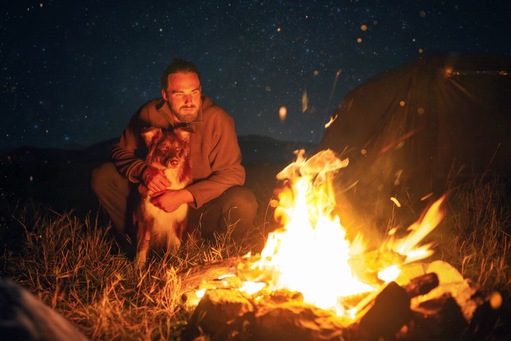 A bearded man in his 20s is sitting behind a bonfire with his dog in front of him.