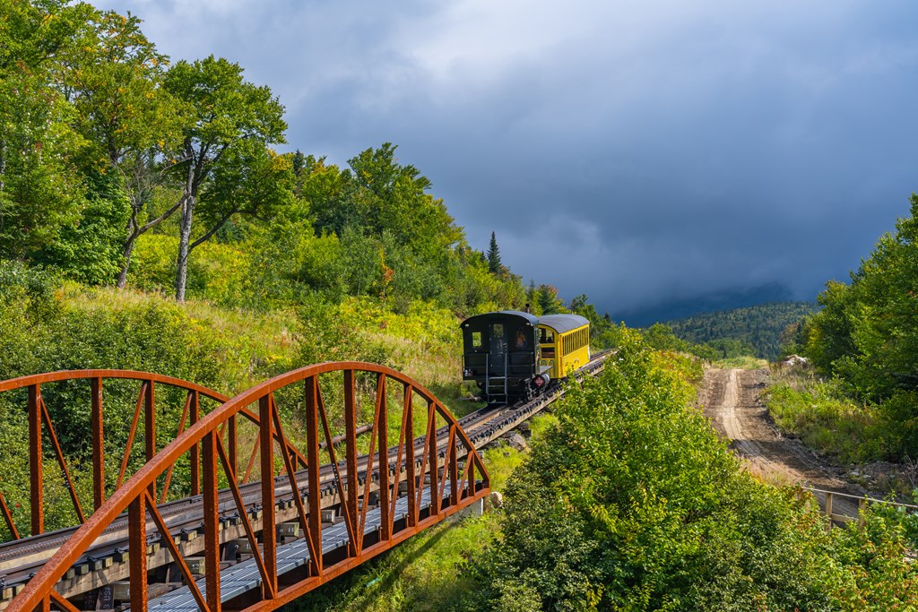 A vintage black locomotive delivers a yellow wagon loaded with tourists on a cog railway to the top of Mount Washington.