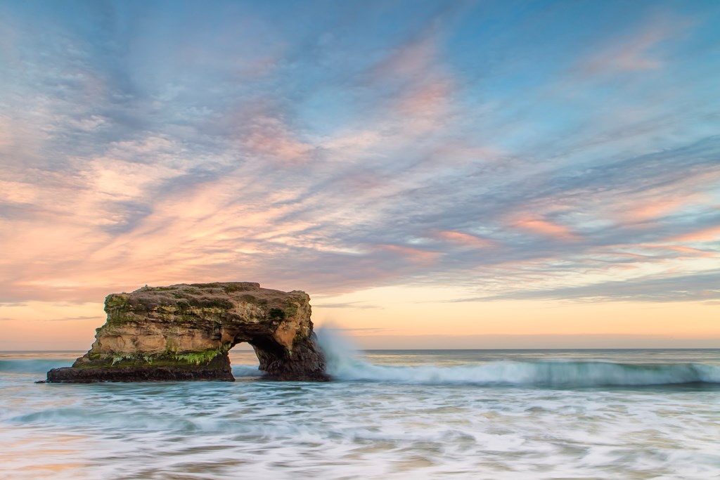 A natural rock formation that looks like a bridge sits in the rising tide during sunset at Natural Bridges State Park, Santa Cruz, California.