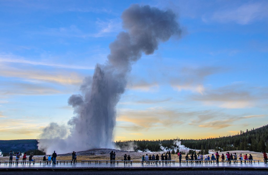 A group of people watch Old Faithful Geyser erupt during sunset in Yellowstone National Park.