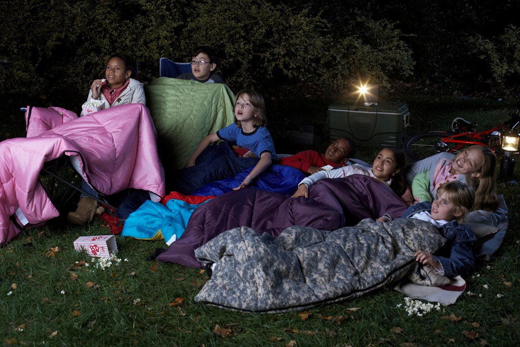 A group of kids in sleeping bags watch a movie outdoors.
