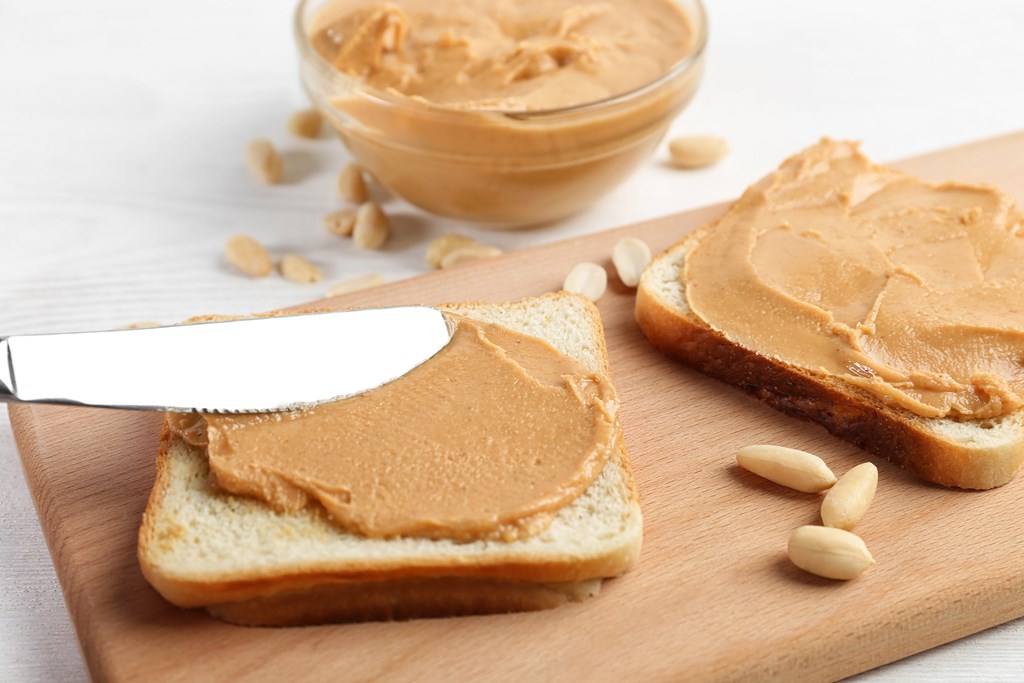 Knife spreading peanut butter on bread on a cutting board.