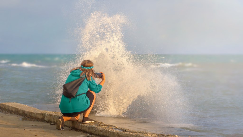 A woman with curly hair, in a green hoodie on a beach flooded with waves takes pictures of a storm on her phone during sunset.
