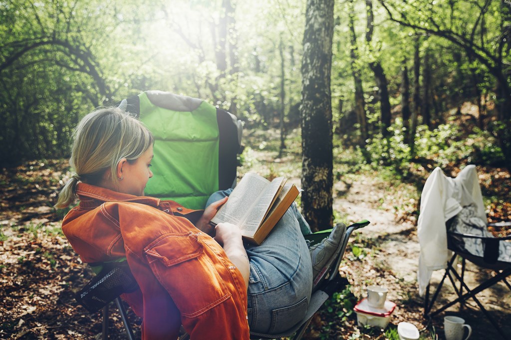 Finding solitude in wilderness concept. Young woman resting in forest, sitting in camp chair and reading book
