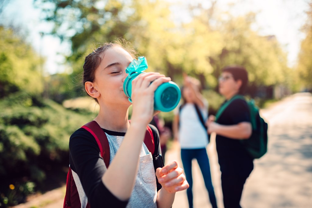 Schoolgirl wearing red backpack drinking water from a reusable water bottle.
