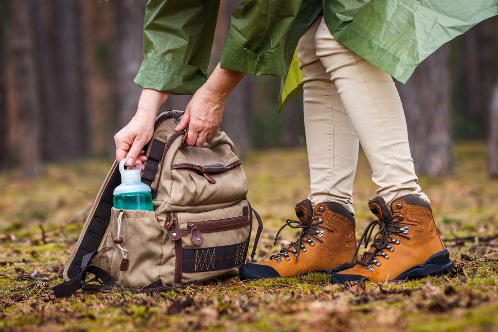 Woman with raincoat and hiking boots taking out water bottle from backpack. 