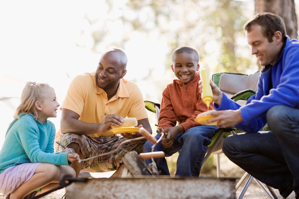 Two fathers with kids roast hot dogs over an open campfire.