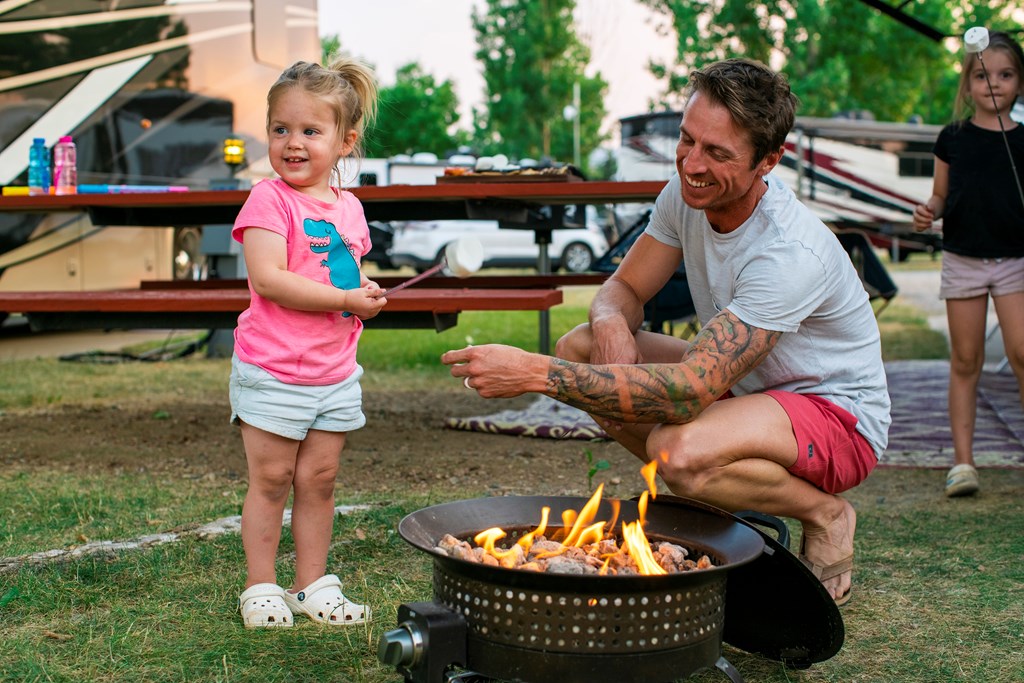A father helps his young daughter roast a marshmallow over a fire.