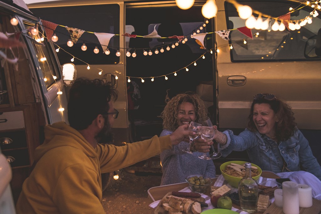 Happy friends cheering and drinking wine during camping vacation at the beach with vintage van.