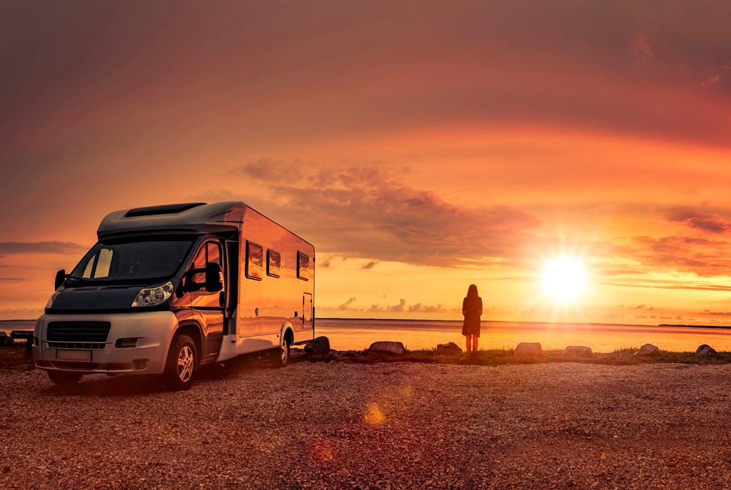 A woman looks out the sunset over the water next to her small motorhome.