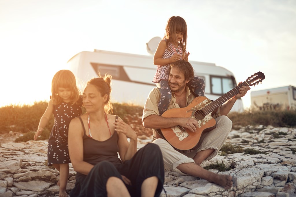 A family of four sits on the beach in front of their RV playing music in the sun.