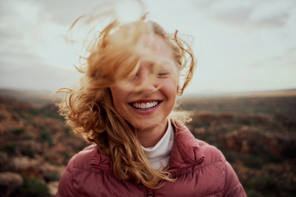 Portrait of young smiling woman face partially covered with flying hair standing at mountain. 