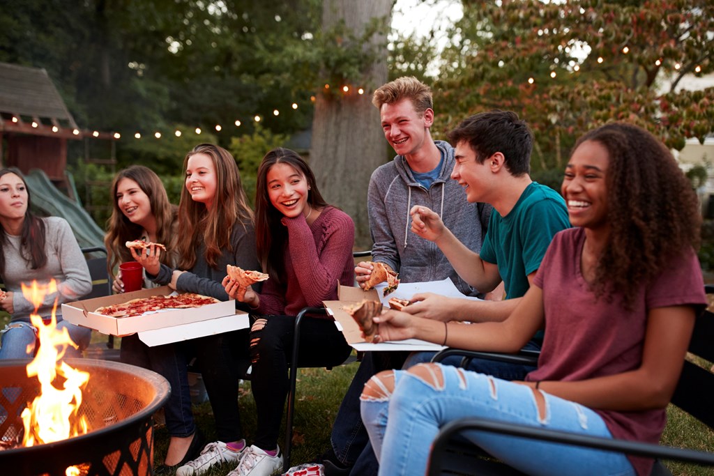 A diverse group of teenagers laughs while eating pizza around a firepit.