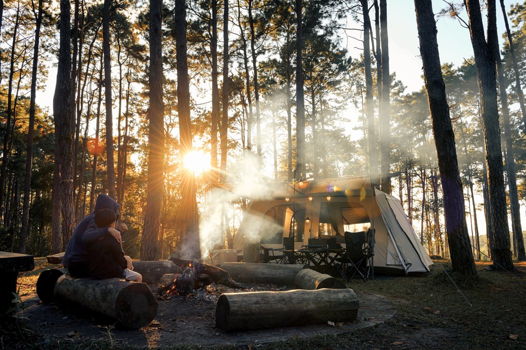 Two people sit by a campfire in front of their tent in a wooded area.