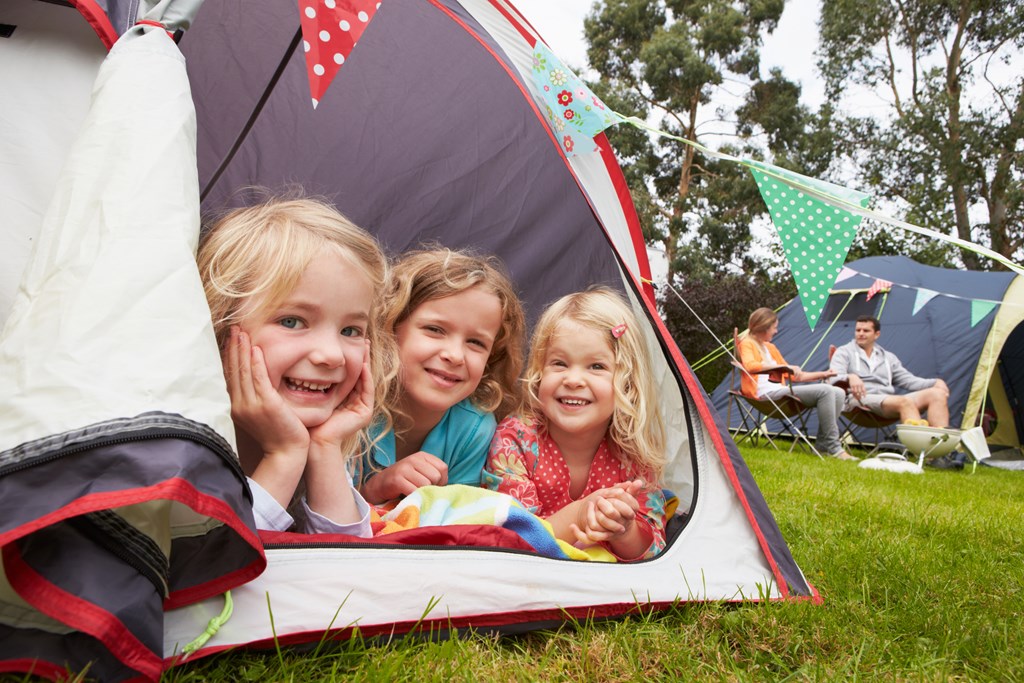 Three young girls smile from the entrance of a camping tent.