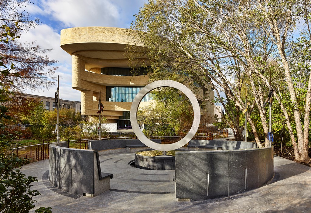 National Native American Veterans Memorial. A stone memorial featuring a vertical ring sits in front of a dramatic building in the modern style.