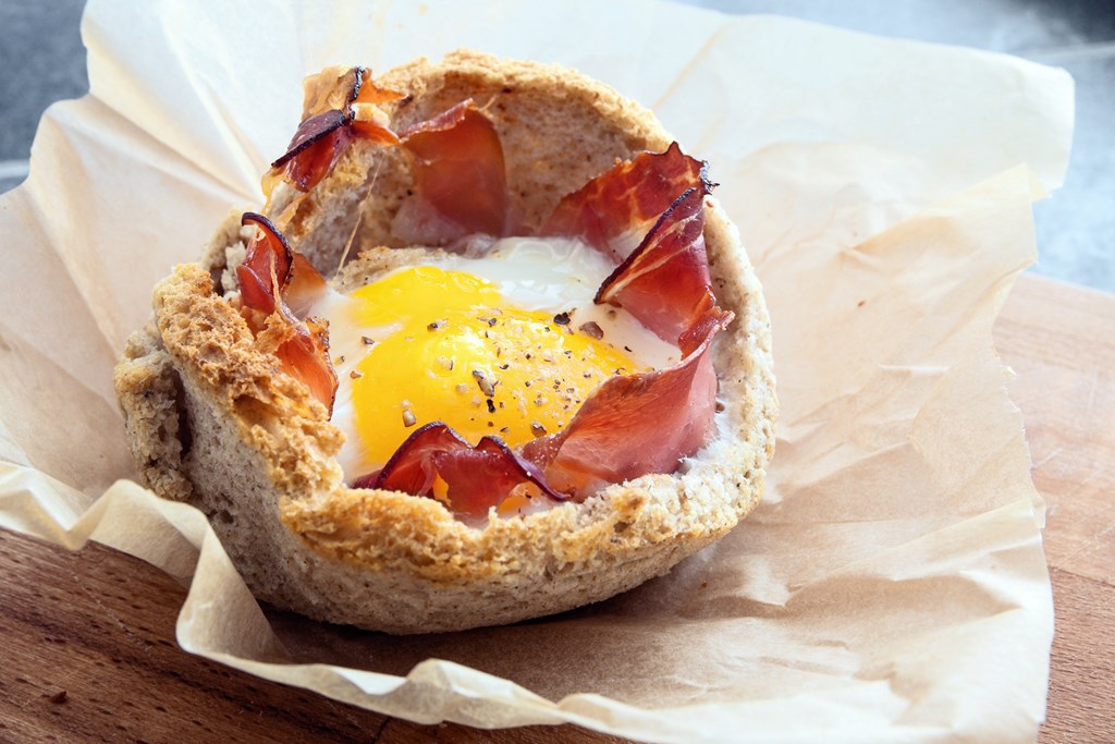 An egg muffin made from toast with bacon and a runny egg in the middle resting on parchment paper.