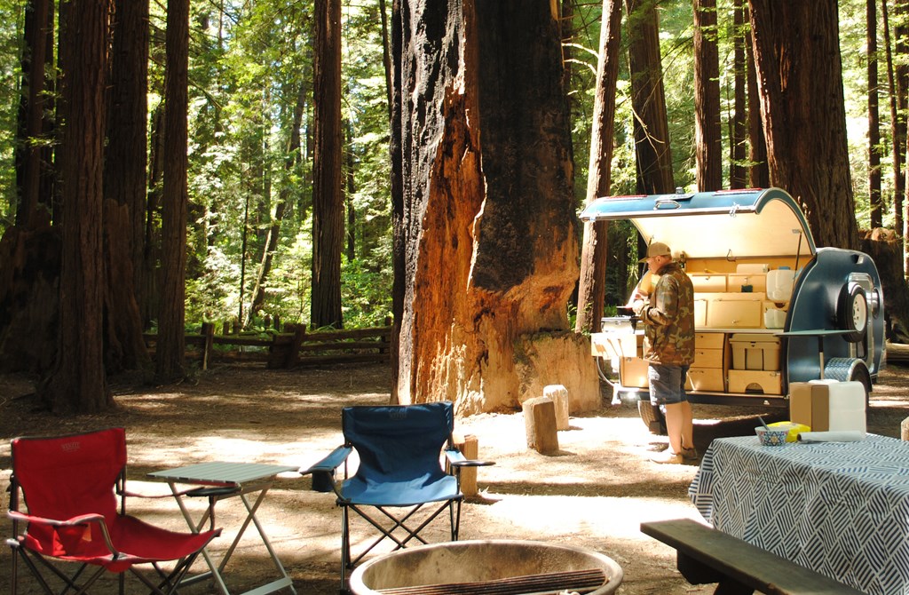 A man cooks over a propane stove in a teardrop trailer kitchen while camping surrounded by redwood trees in Humboldt Redwoods State Park in California.