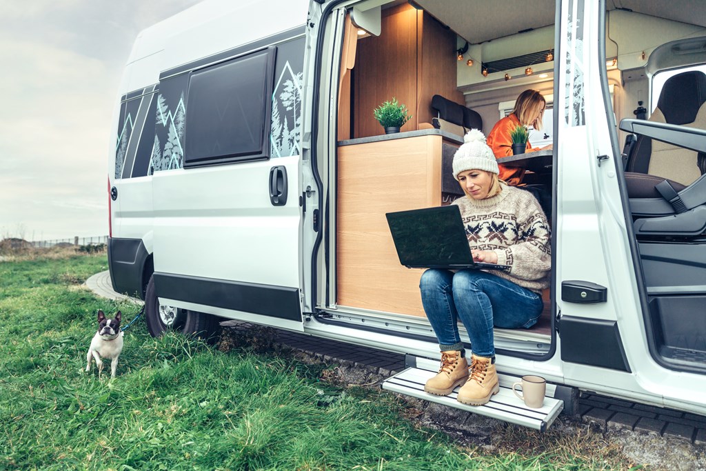 Woman teleworking sitting in the door of a camper van while her partner works inside.