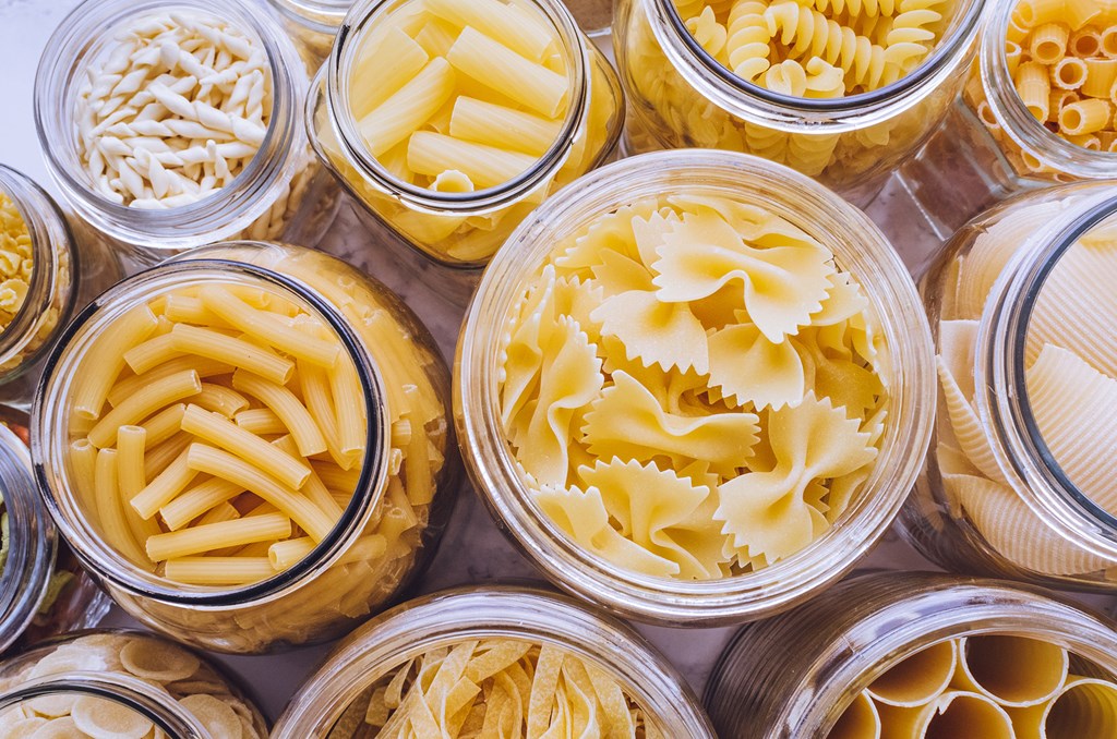 Variety of types and shapes of Italian pasta in glass jars on marble background.