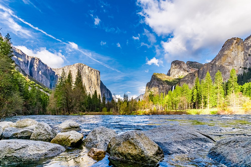 Landscape image of El Capitan and Half Dome in Yosemite National Park with the rocky shore of the Merced River in the foreground, with a vibrant blue cloudy sky.