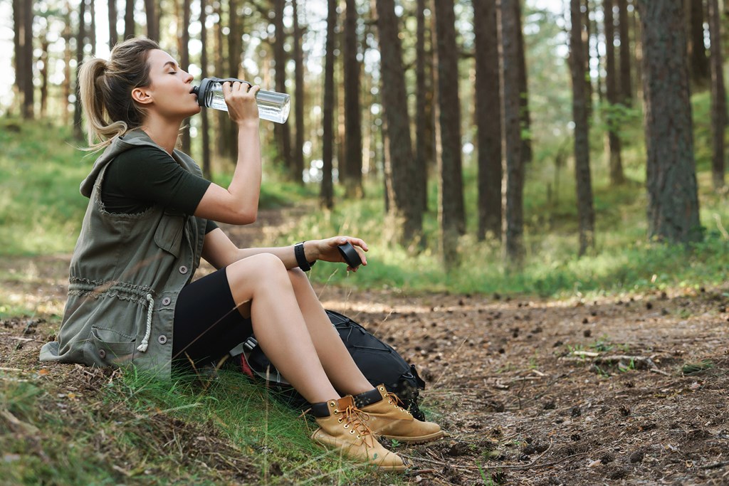 Young female hiker drinking water from reusable bottle in green forest