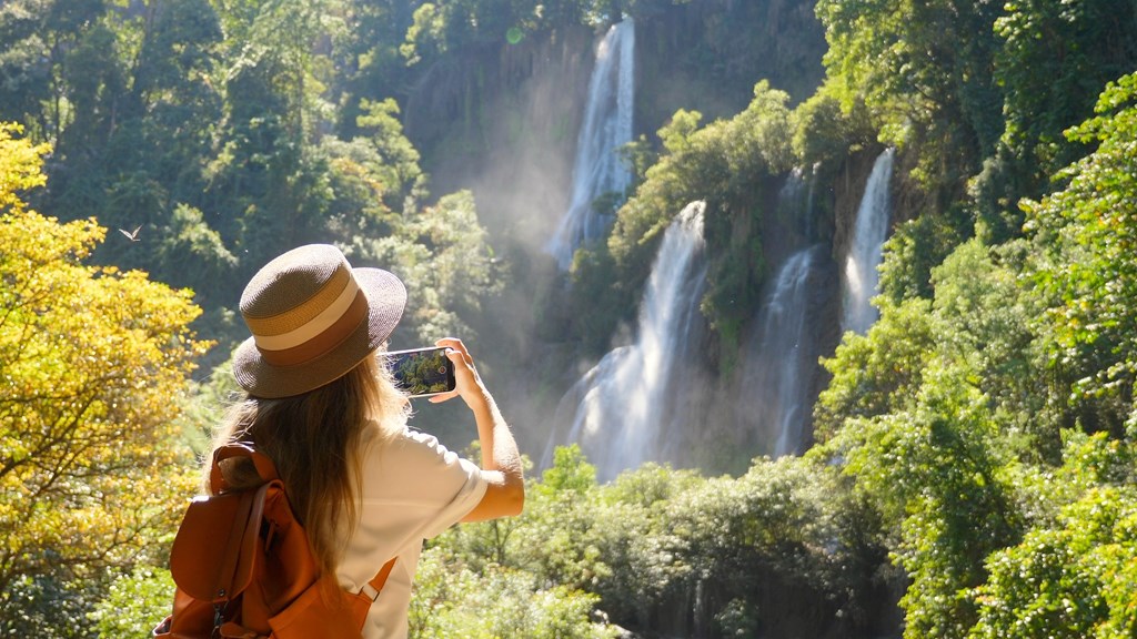 Back view of millennial travel woman on trip or adventure. Young travel blogger on waterfall, make photos on smartphone for social media. Amazing landscape of Thi Lor Su waterfall in Thailand