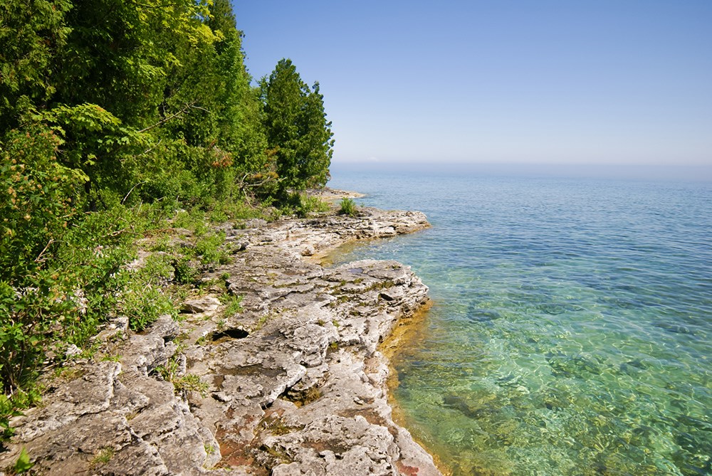 Bright blue lake to the right, gray rocky terrain with green trees on the left in Peninsula State Park in Door County Wisconsin